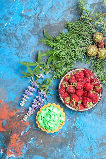 Top view of berry bowl, small tart and tree branches on blue surface