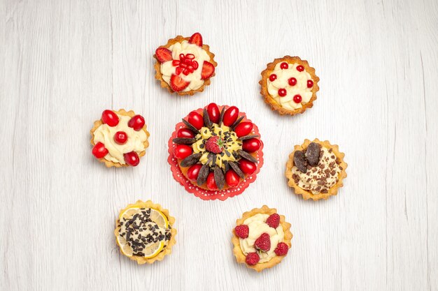 Top view berry cake on the red oval lace doily and tarts on the white wooden ground