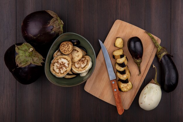 Top view black eggplant with wedges in a bowl and on a cutting board with a knife on a wooden background