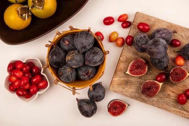 Top view of black figs on a bucket with cornelian cherries on a bowl with black figs and cornelian cherries isolated on a wooden kitchen board on a white wall