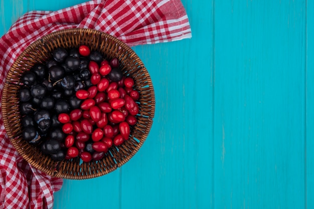 Free photo top view of black grape on a bucket with red cornel cherries on a bucket on a red checked cloth on a blue wooden background with copy space