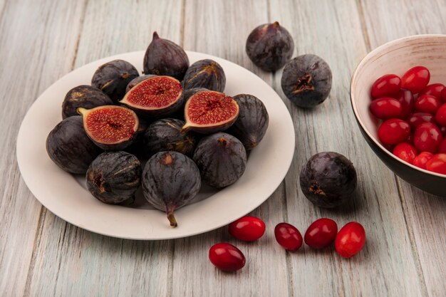 Top view of black mission figs on a white bowl with cornelian cherries on a grey wooden surface