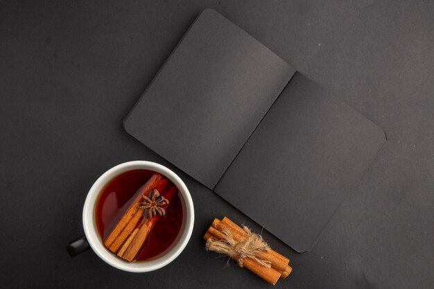 Top view black notepad cup of tea flavored by cinnamon and anise cinnamon sticks tied with rope on dark table