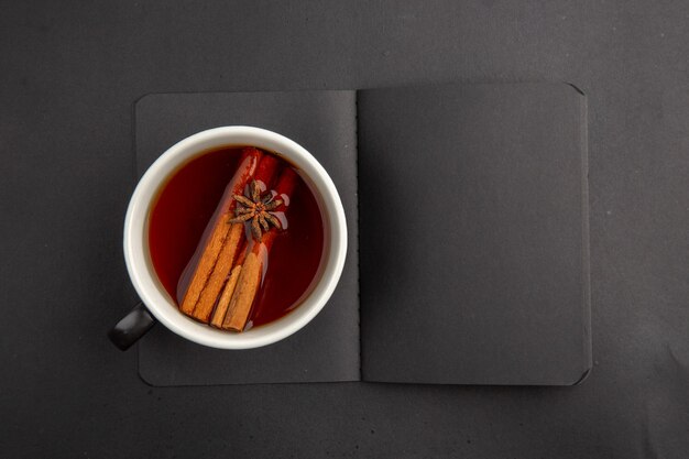 Top view black notepad cup of tea flavored by cinnamon and anise on dark table