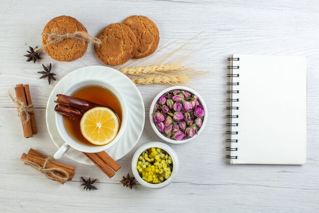 Top view of black tea with cinnamon lime lemon and various herbals cookies next to notebook on white background