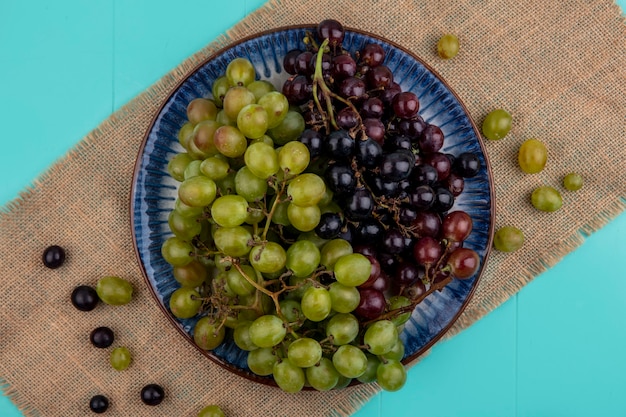 Top view of black and white grapes in plate on sackcloth on blue background