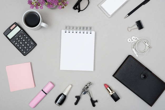 Top view of blank notepad surrounded by coffee cup; calculator; make-up objects and earphone on gray background