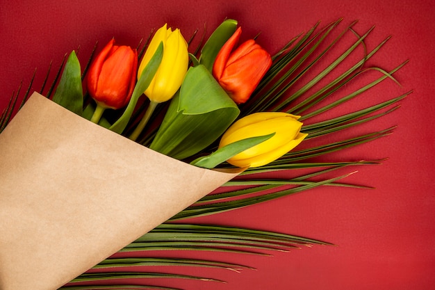 Free photo top view of a bouquet of yellow and red color tulips in craft paper with palm leaf on red table