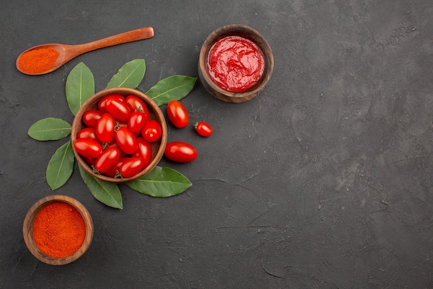 Free photo top view a bowl of cherry tomatoes bay leaves a wooden spoon and bowls of ketchup and hot pepper powder on the black table