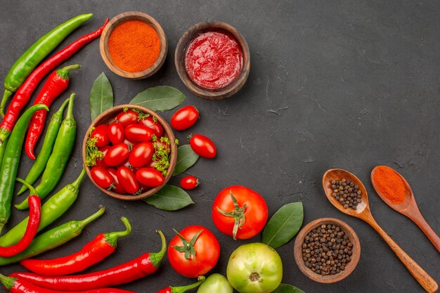 Top view a bowl of cherry tomatoes hot red and green peppers bay leaves spices in wooden spoons bowls of ketchup hot red pepper powder and black pepper and tomato on black