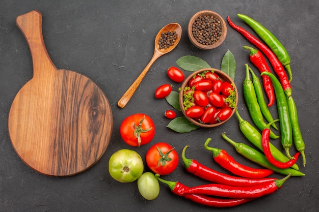 Top view a bowl of cherry tomatoes hot red peppers black pepper in a wooden spoon a bowl of black pepper and an oval chopping board on black background