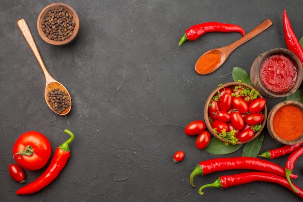 Top view a bowl of cherry tomatoes hot red peppers black pepper in a wooden spoon bowls of ketchup black pepper and red pepper powder on the black table