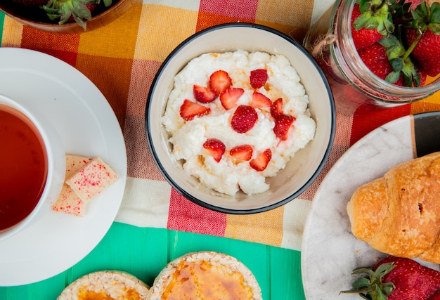 Top view of bowl of cottage cheese with strawberries cup of tea crispbreads crescent roll on cloth on green surface