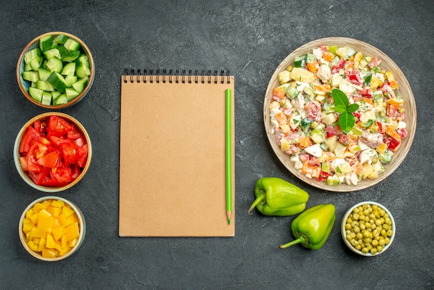 Top view of bowl of vegetable salad with bowls of vegetables notepad and bell peppers on side on dark green table