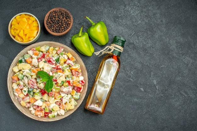 Top view of bowl of vegetable salad with bowls of vegetables and pepper oil bottle and bell peppers on side on dark background