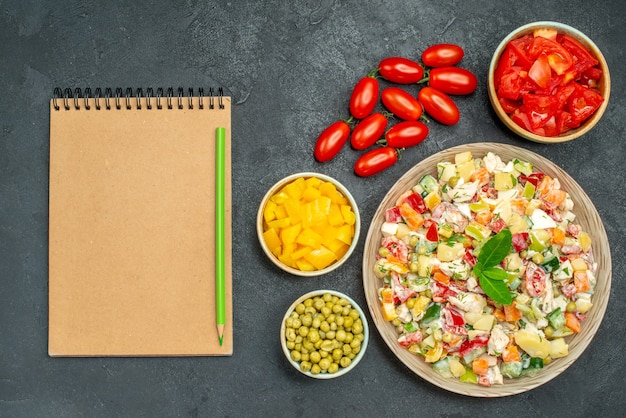 Top view of bowl of vegetable salad with vegetables and notepad on side on dark grey background