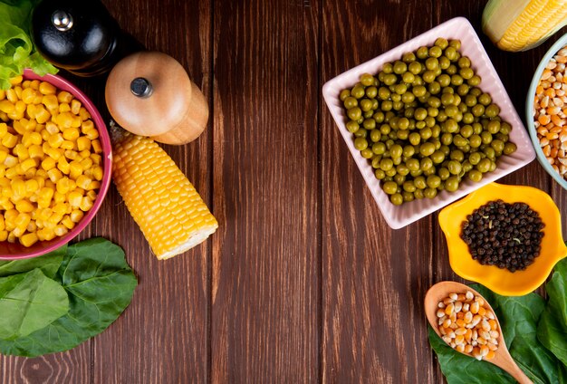 Top view of bowls of green peas corn seeds and black pepper with spinach on wooden surface