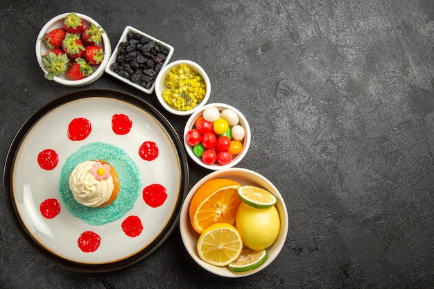 Top view bowls of sweets bowls of different sweets citrus fruits and berries next to the plate of cake with cream and sauces on the black table