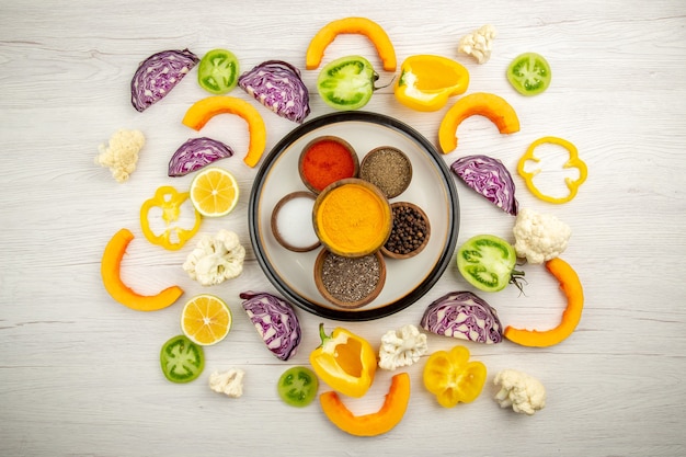 Top view bowls with spices on round plate turmeric salt black pepper red pepper chopped vegetables on white surface