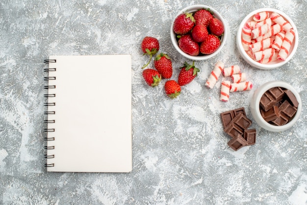 Top view bowls with strawberries chocolates candies and some strawberries chocolates candies at the right side and a notebook at the left side of the grey-white table