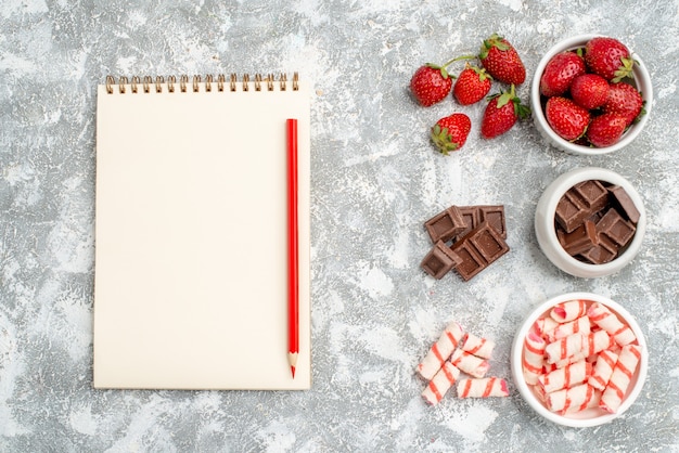 Top view bowls with strawberries chocolates candies and some strawberries chocolates candies at the right side and notebook with red pencilat the left side of the grey-white ground