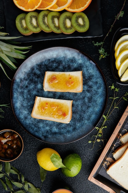 Top view of bread slices smeared with jam with citrus fruits around as pineapple kiwi orange lime lemon and tangerine slices with lemon bread slices in tray on black background