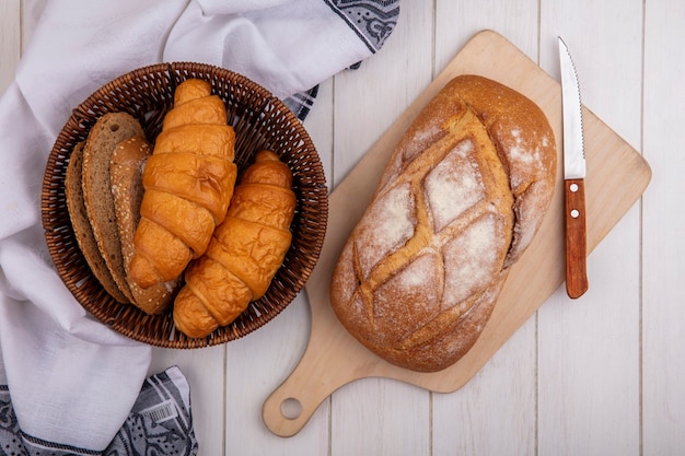 Top view of breads as croissant and seeded brown cob bread slices in basket on cloth and crusty bread on cutting board on wooden background