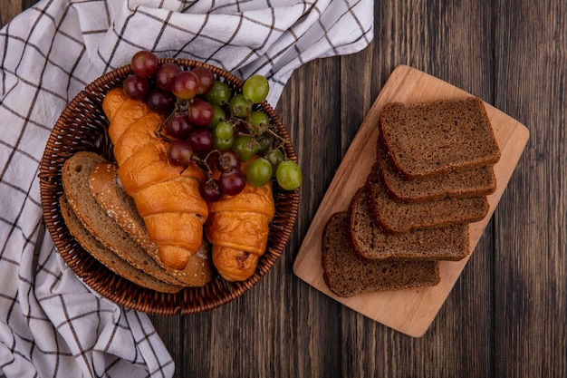 Top view of breads as croissants and seeded brown cob bread slices with grape in basket on plaid cloth and rye bread slices on cutting board on wooden background