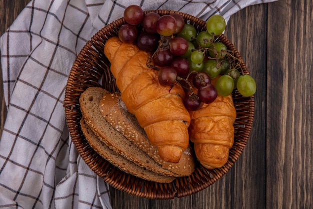 Top view of breads as croissants and seeded brown cob bread slices with grape in basket on plaid cloth on wooden background