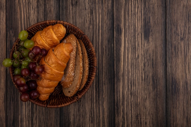 Top view of breads as croissants and seeded brown cob bread slices with grape in basket on wooden background with copy space