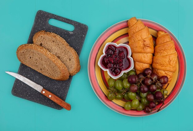 Top view of breads as seeded brown cob with knife on cutting board and croissants with grapes and raspberry jam in plate on blue background