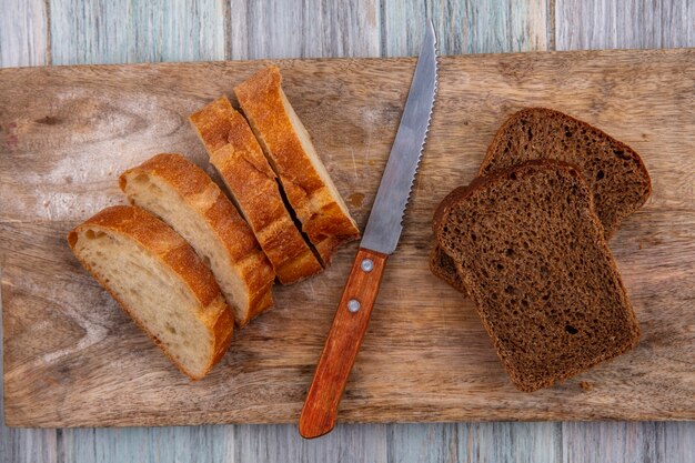 Top view of breads as sliced baguette and rye ones with knife on cutting board on wooden background