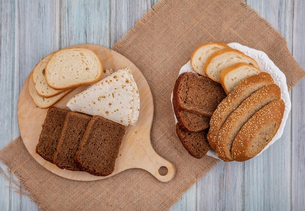 Top view of breads as sliced rye white seeded brown cob ones and flatbread on cutting board and in plate on sackcloth on wooden background