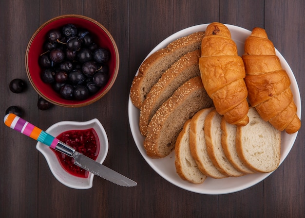 Top view of breads as sliced seeded brown cob baguette and croissants in plate and bowls of raspberry jam and sloe berries with knife on wooden background