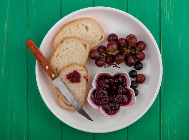 Top view of breakfast set with bread slices raspberry jam and grape with knife in plate on green background