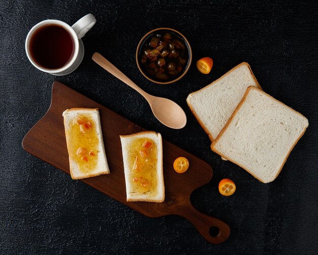 Top view of breakfast set with jam smeared on bread slices on cutting board with jam bread slices cup of tea kumquats and spoon on black background