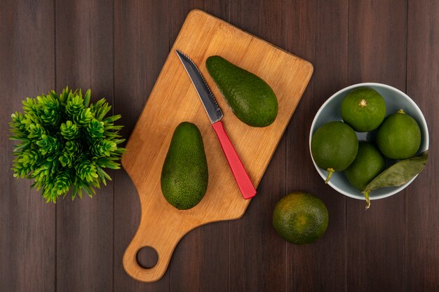 Top view of bright green avocados on a wooden kitchen board with knife with limes on a bowl with tangerine isolated on a wooden background