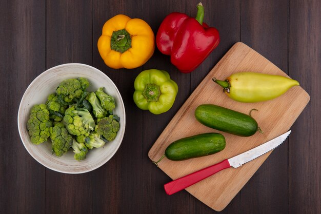 Top view broccoli in bowl with bell peppers  cucumbers on cutting board with knife  on wooden background