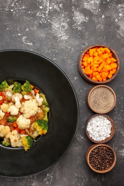 Top view broccoli and cauliflower salad in black bowl different spices and cutting carrot in bowls on dark surface free place