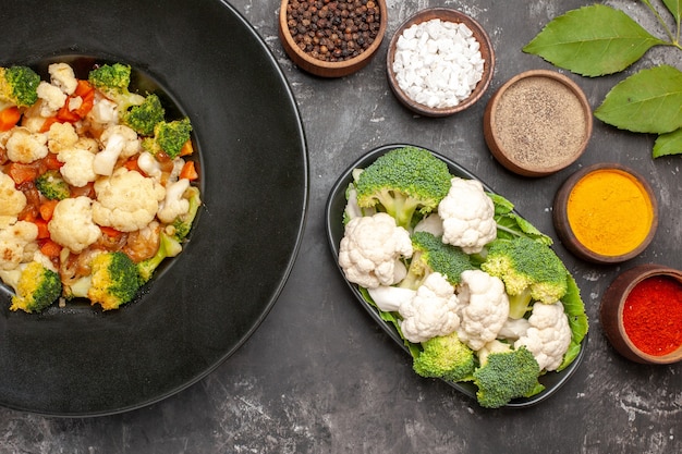 Top view broccoli and cauliflower salad in black bowl raw broccoli and cauliflower on plate different spices on dark surface