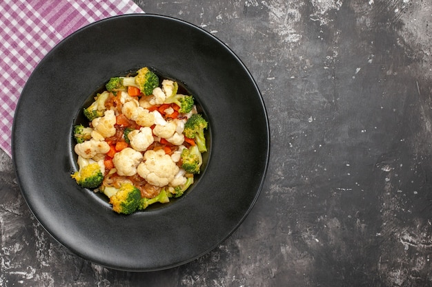 Top view broccoli and cauliflower salad on black oval plate pink and white checkered tablecloth on dark surface with free space