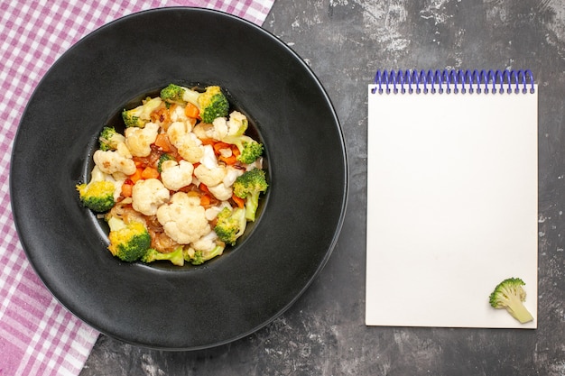 Top view broccoli and cauliflower salad on black oval plate pink and white checkered tablecloth a notebook on dark surface