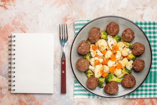 Top view broccoli and cauliflower salad and meatball on plate green and white checkered napkin a fork a notebook on nude isolated background