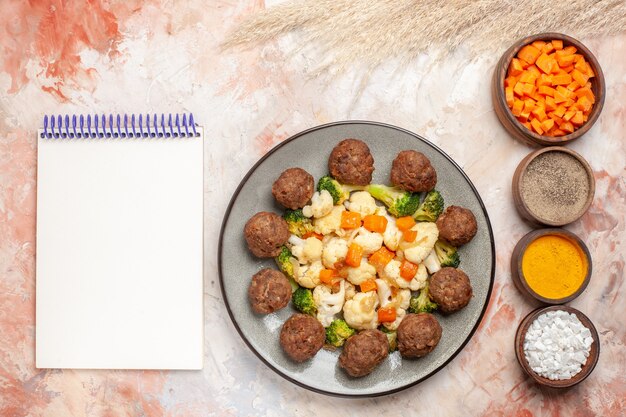 Top view broccoli and cauliflower salad and meatball on white plate vertical row bowls with different spices a notepad on nude surface