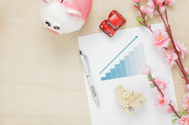 Free photo top view business office desk concept. saving money with charted summary  wood house  also car and clock on wooden shelf.the beautiful pink flower with pen on wood background with copy space.