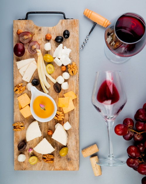 Free photo top view of butter with different kinds of cheese grape olives nuts on cutting board and glasses of wine with corks and corkscrew on white