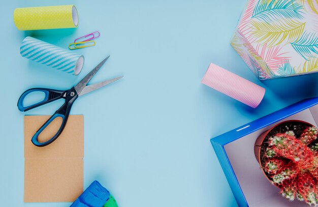 Top view of a cactus in a flowerpot in a carton gift box with scissors colorful paper clips and rolls of adhesive tape on blue background