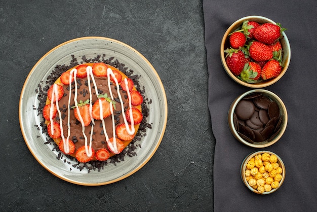 Top view cake bowls of hazelnut strawberry and chocolate on tablecloth and cake with chocolate and strawberry on black table