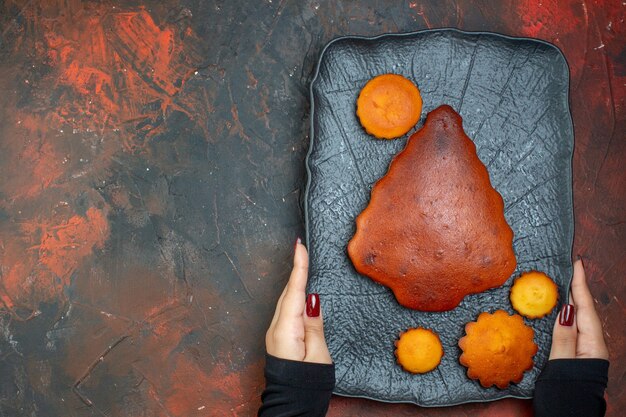 Top view cake and cupcakes on black plate in female hands on dark red table with free place