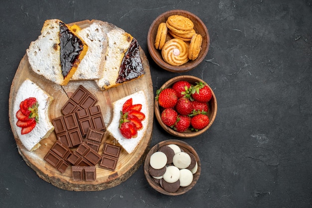 Top view cake slices with fruits cookies and choco bars on dark surface